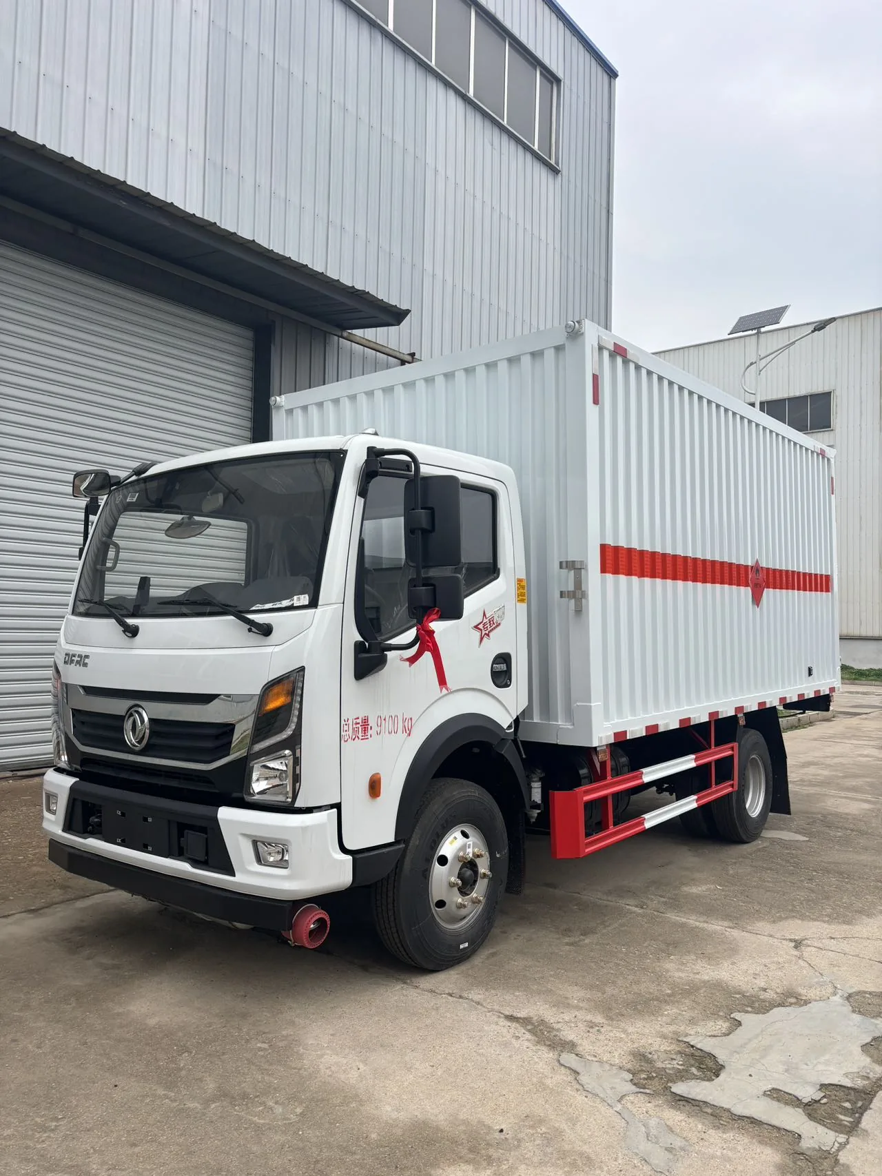 东风多利卡 D6 黄牌 5 吨危险品厢式运输车，前左 45 度视角，瓦楞板白厢体（垂直条纹板工艺）+ 「总质量 9100kg」红字 + 驾驶室左后视镜系红色丝带（刚下线喜庆场景），背景为厂区卷闸门与路灯