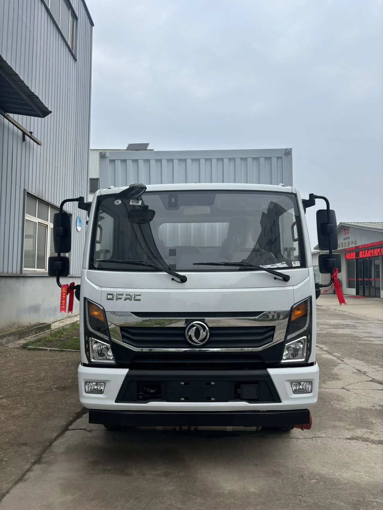 东风多利卡 D6 黄牌 5 吨危险品厢式运输车，正脸驾驶室特写，白色 DFAC 驾驶室 + 圆形双飞燕东风 logo + 镀铬中网 + 矩形大灯 + 左右大视野后视镜 + 后视镜系红色飘带，背景为厂区建筑