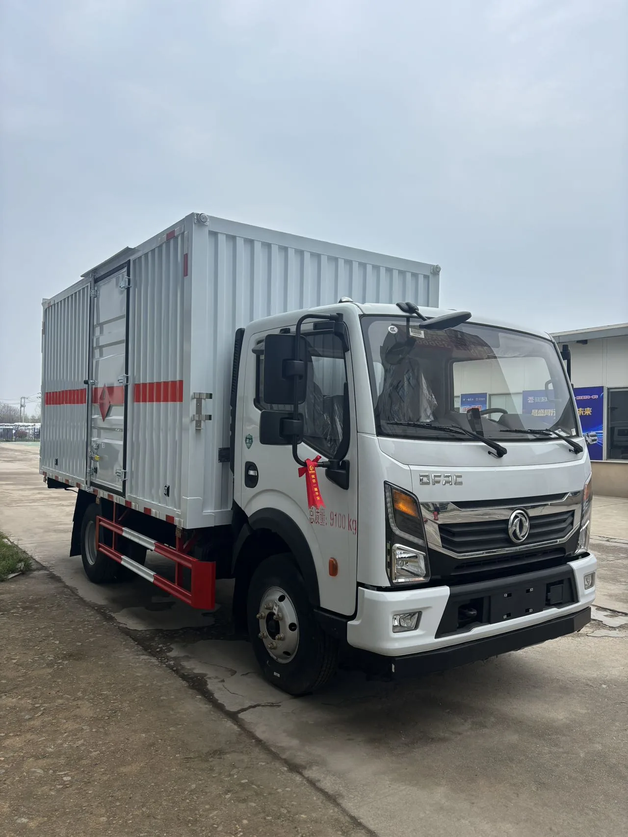 东风多利卡 D6 黄牌 5 吨危险品厢式运输车，前左 45 度视角，瓦楞板白厢体 + 红色防护栏 + 「总质量 9100kg」红字 + 左侧后视镜系红色飘带（刚下线喜庆场景），背景为蓝色 LED 招牌
