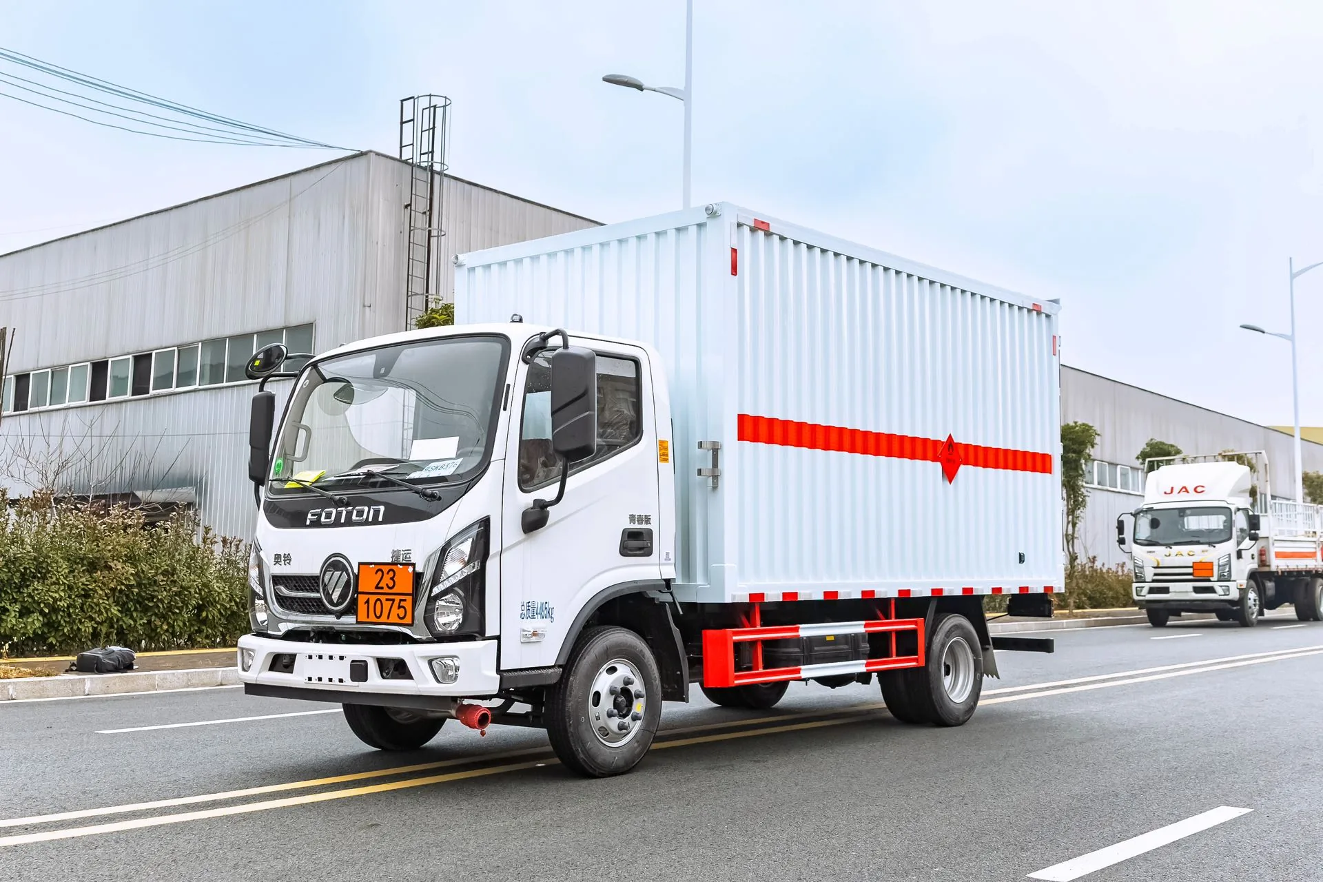  福田奥铃蓝牌 4.2 米危险品厢式运输车，右前 45 度视角，瓦楞结构厢体（垂直条纹板）+ 危险品菱形标识 + 橙色反光横条 + 红色防护栏，黄色车牌 23·1075，驾驶室门标 FOTON 标志，背景为厂区建筑与江淮厢式车