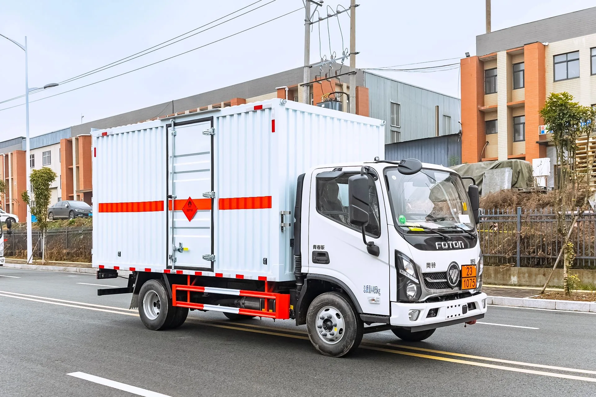  福田奥铃蓝牌 4.2 米危险品厢式运输车，后右 45 度视角，瓦楞结构厢体（垂直条纹板）+ 后门单开门设计 + 危险品菱形标识 + 橙色反光横条，黄色车牌 23·1075，驾驶室标 FOTON 标志，背景为商贸城建筑与电缆塔