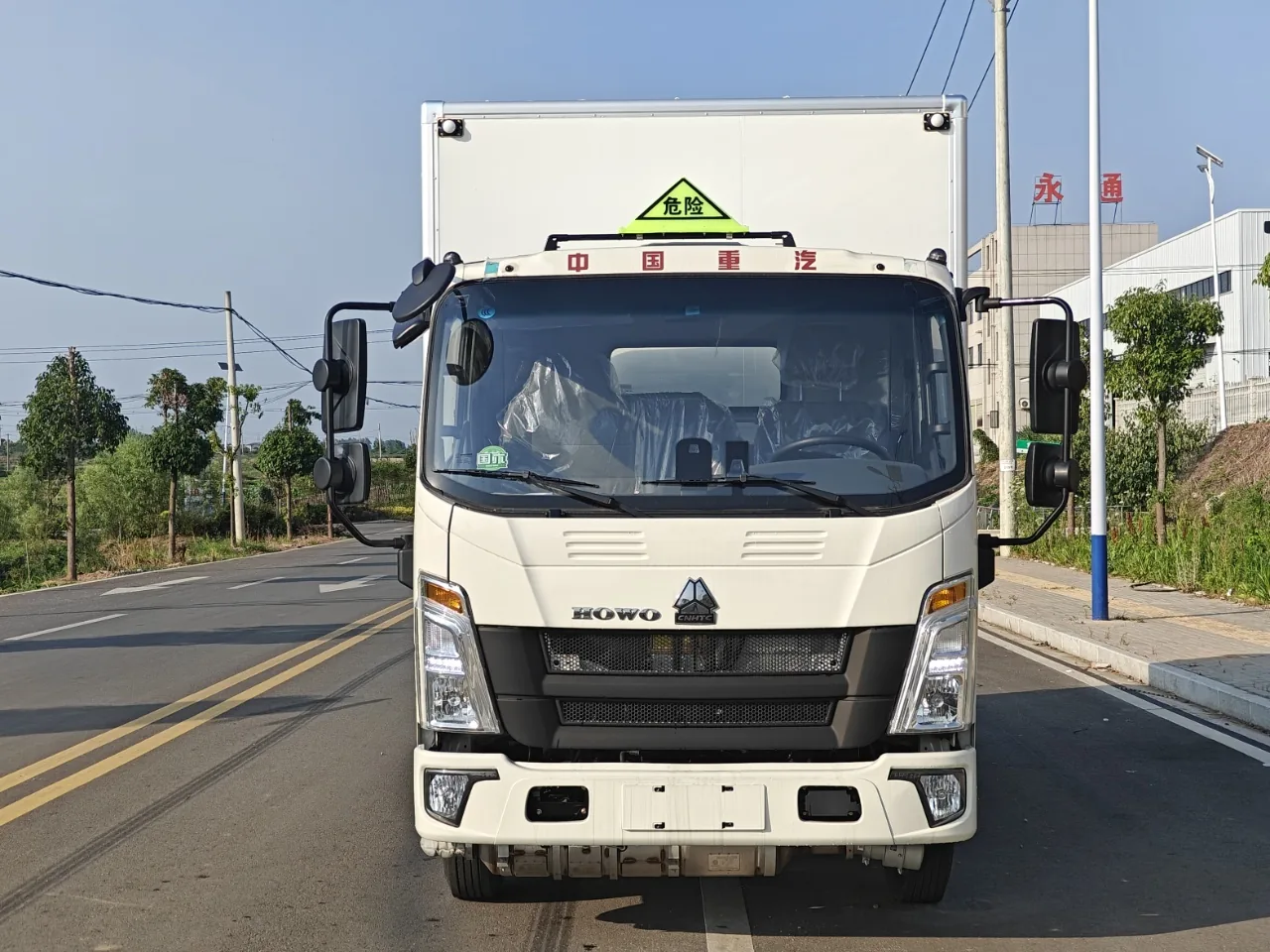 重汽豪沃蓝牌危险品厢式运输车 车头正视图，白色驾驶室，前挡风带「危险」三角顶灯与厂区场景