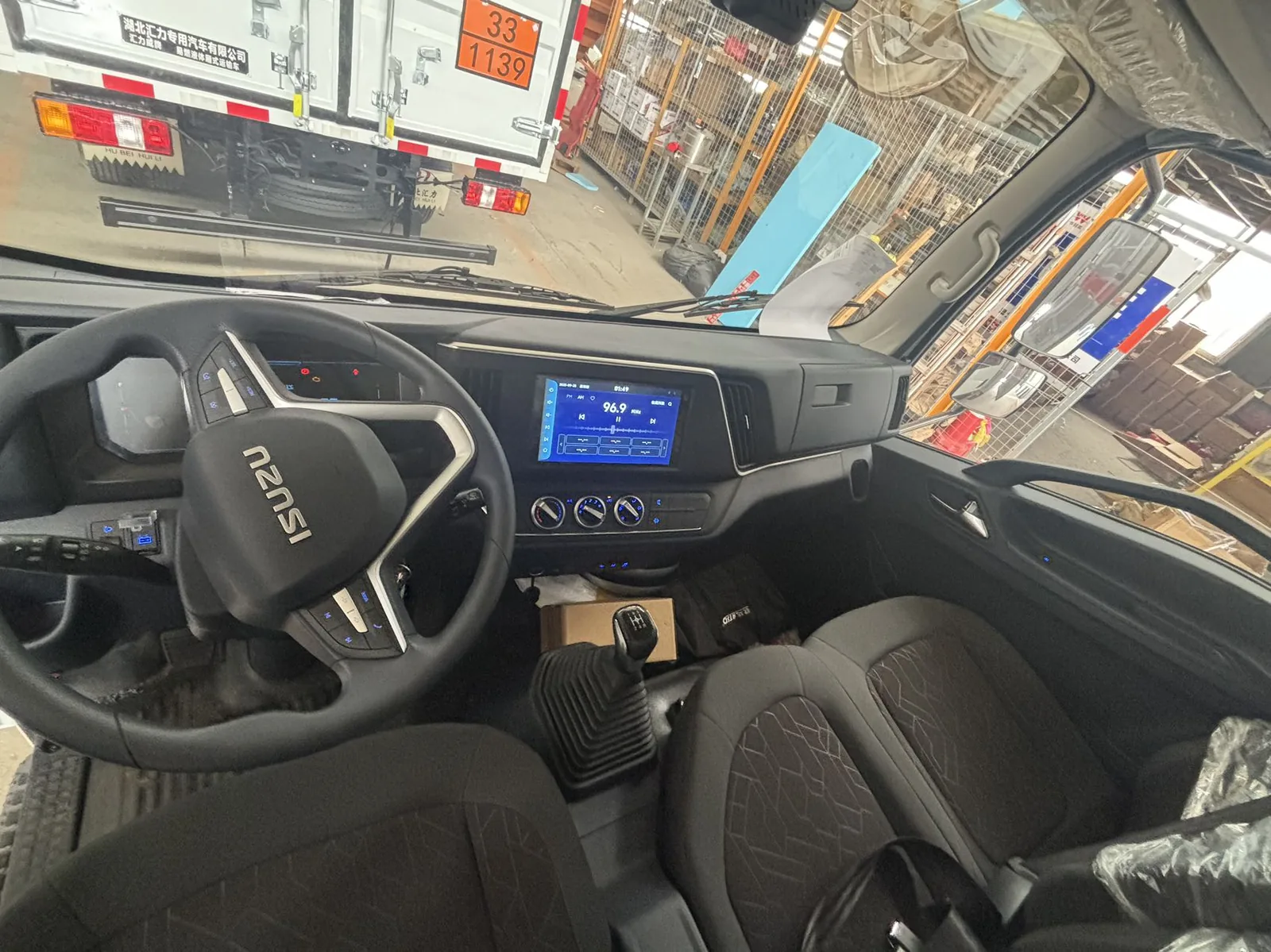 江西五十铃蓝牌危险品厢式运输车驾驶室内部实拍，ISUZU 多功能方向盘（多功能按键）+ 中控大屏（倒车影像界面）+ 手动挡杆 + 双人座椅 + 空调旋钮，车间内拍摄