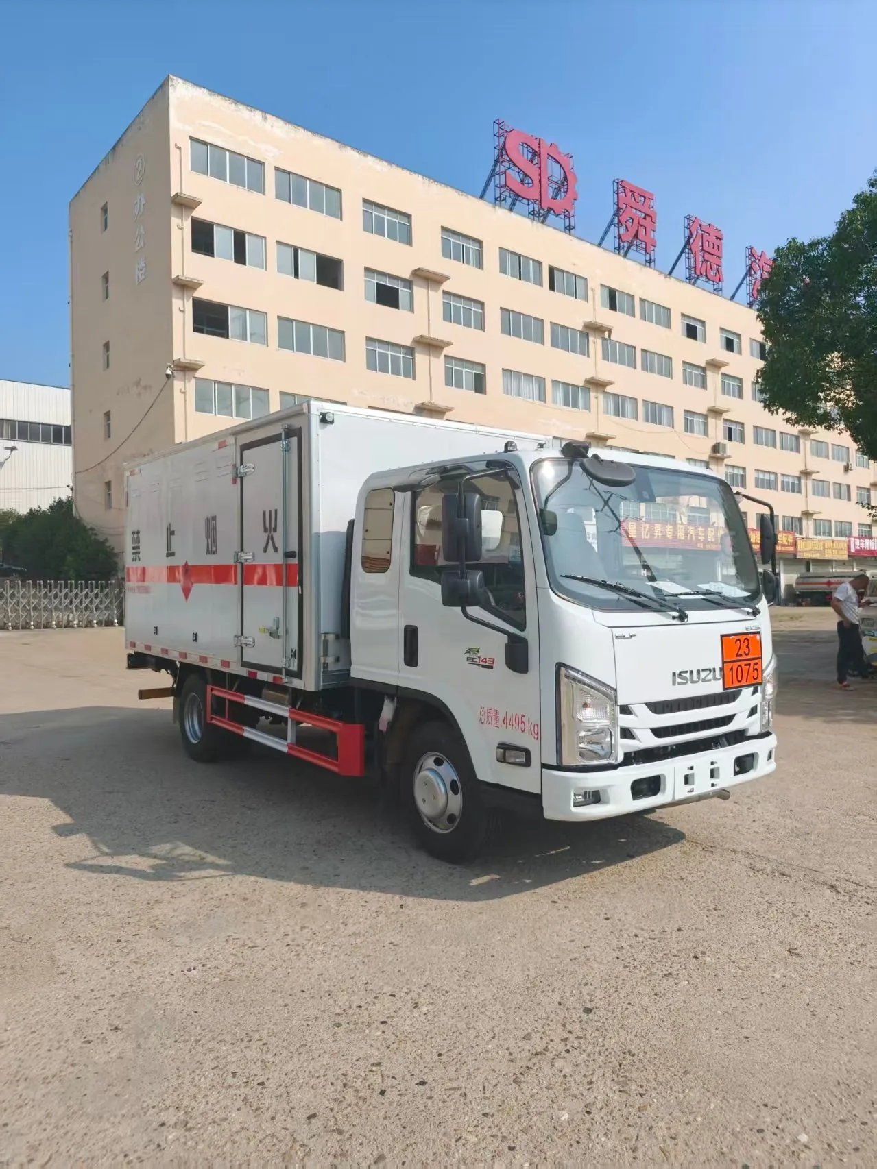 江西五十铃蓝牌危险品厢式运输车实拍，右前 45 度视角，白色 ISUZU 驾驶室（143 马力标）+ 白色厢体（「禁止烟火」字样 + 橙色反光带 + 危险品菱形标志牌）+ 侧门 + 总质量 4495kg 喷字 + 7.00R16 轮胎，背景为舜德汽车大楼