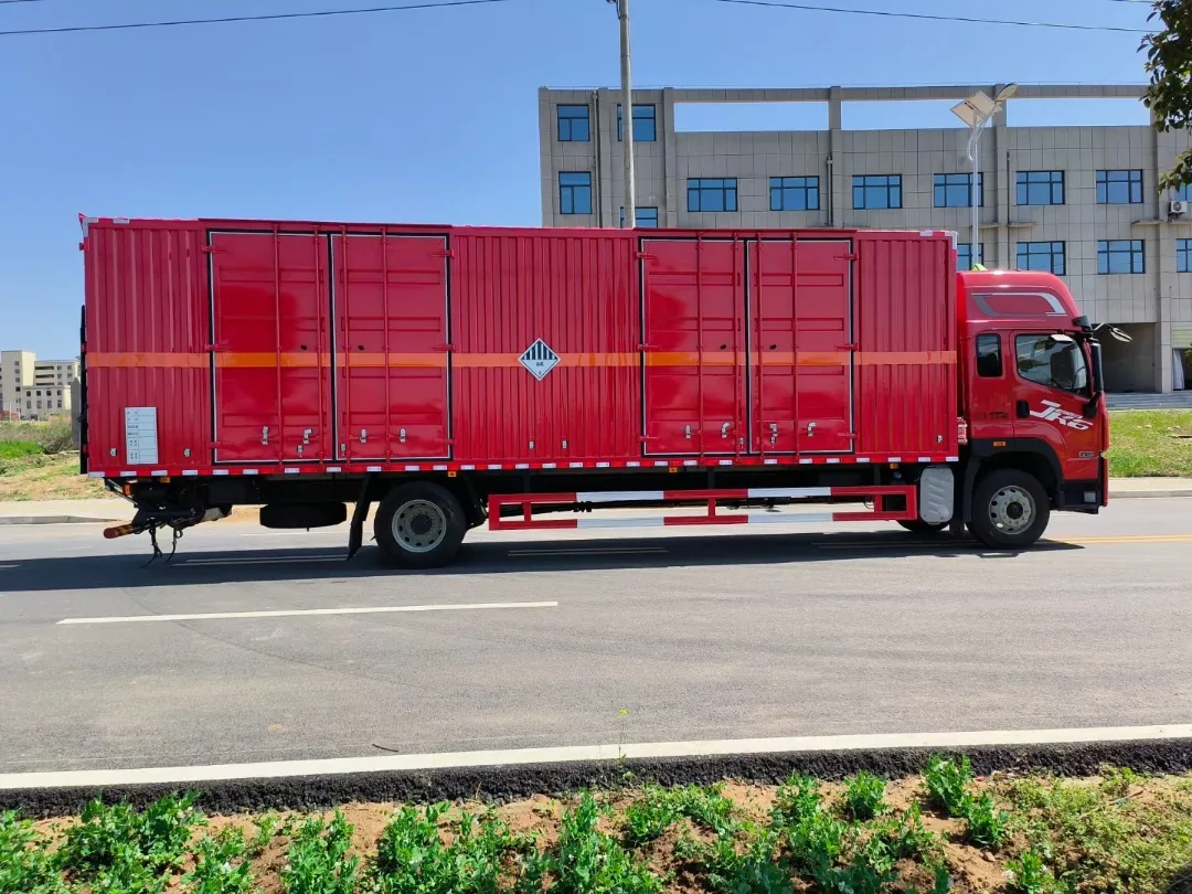 解放JK6单桥杂项危险品厢式运输车 右侧正侧面实拍：红色瓦楞厢体带两个侧开门 + 杂项危险物品菱形标志 + 橙色反光腰线，厂房背景