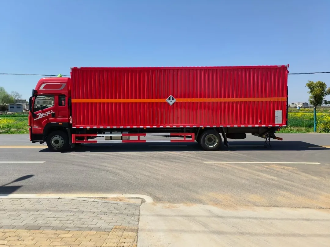 解放JK6单桥9.6米危险品厢式运输车 左侧正侧面实拍：红色瓦楞厢体 + 橙色反光大带 + 杂项危险物品菱形标 + 435升级冲焊桥，乡村公路背景