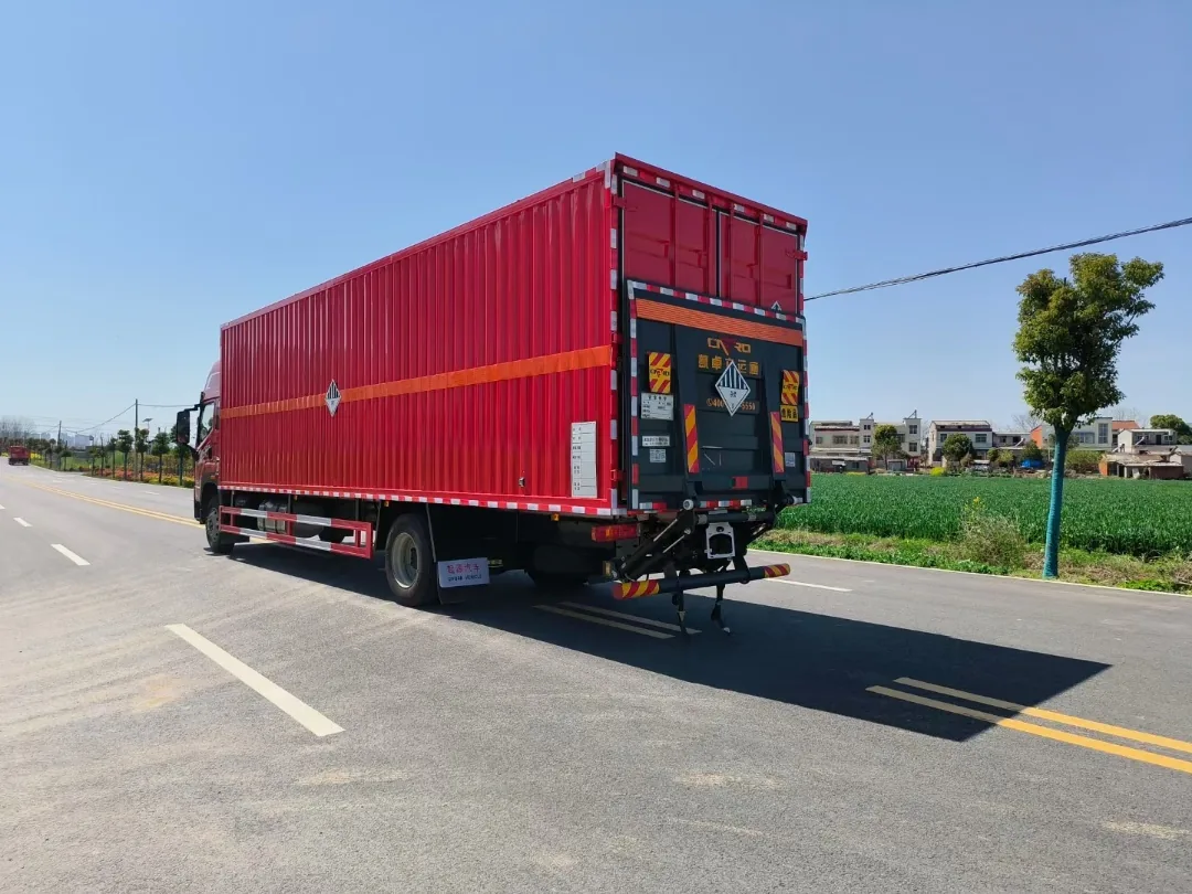 解放JK6单桥危险品厢式运输车 左后45度实拍：尾部液压升降尾板 + 红色瓦楞厢体 + 杂项危险物品菱形标志，乡村公路
