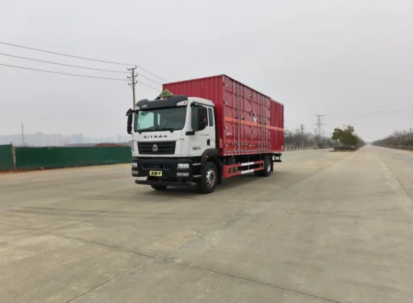 众德牌 重汽汕德卡 9.6 米危险品厢式运输车，左前 45 度视角，SITRAK 标志在驾驶室前面（品牌正面最清晰）+ 白色驾驶室 + 红色厢体（瓦楞结构+橙色反光带+菱形告示牌）+ 红色防护栏 + 危险品三角顶灯 + 绿色施工围挡背景，厂区外公路