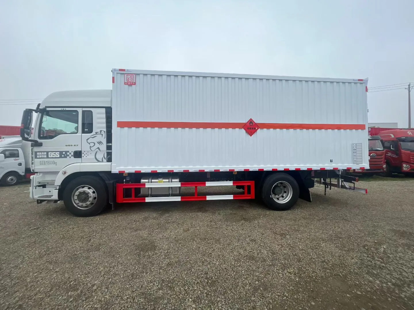 众德牌 重汽汕德卡 G5S 9.6 米危险品厢式运输车，左正侧视角，白色厢体（瓦楞结构 + 红色腰线 + 9 类菱形告示牌 + 红色反光带）+ 红色防护栏 + 295/80R22.5 重载胎 + 白色驾驶室（SITRAK 标志 + G5S 字样），厂区停车场，背景为多辆红色重型卡车